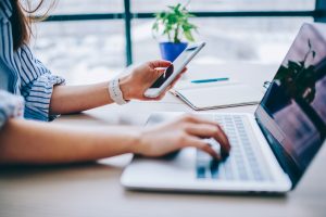 Person at a desk with a mobile phone in hand while also typing into a laptop